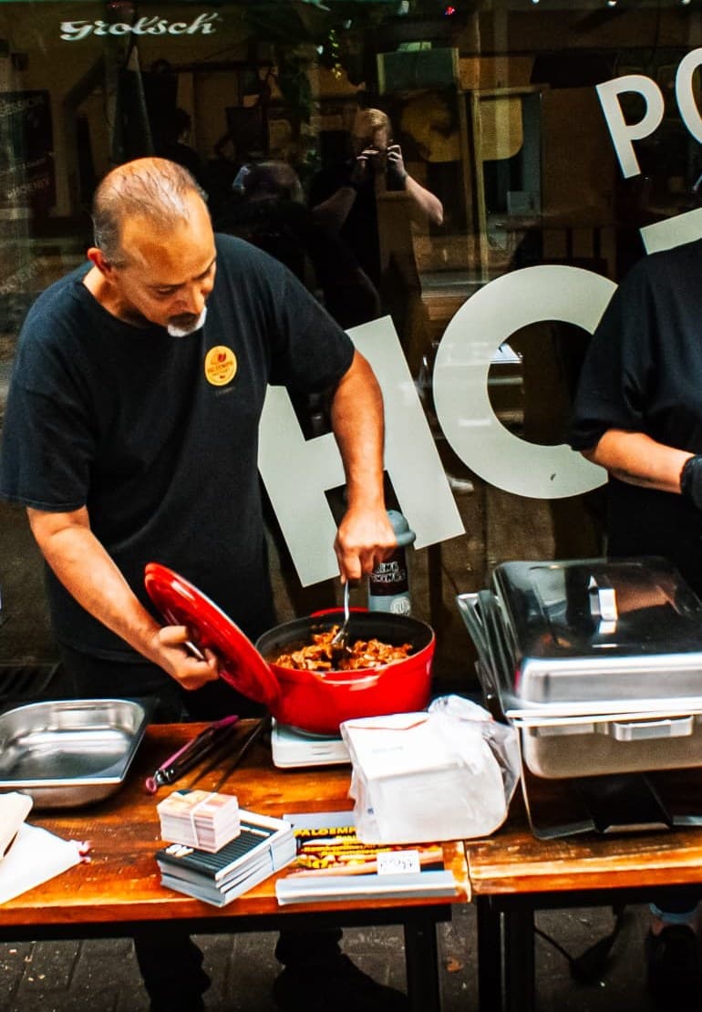 Dennis licht het deksel van een pan ketjapkip op zijn eigen kraam op de Bricks Blockparty in Breda, in shirt met het logo van Paloempie Factory