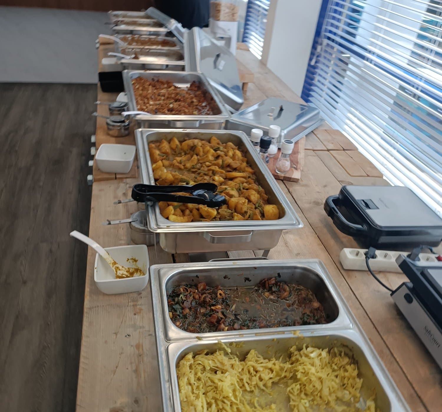 Een rij chafing dishes op een houten tafel met aardappelmassala, gestoofd bladgroen en noedels, op locatie bij een opdracht